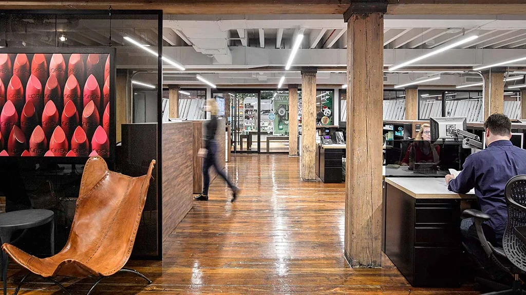 An industrial-style office hallway with wood columns and exposed ceiling beams, a leather butterfly chair and lipstick-patterned artwork in the foreground, a person walking past glass-walled offices, and an employee working at a desk on the right.
