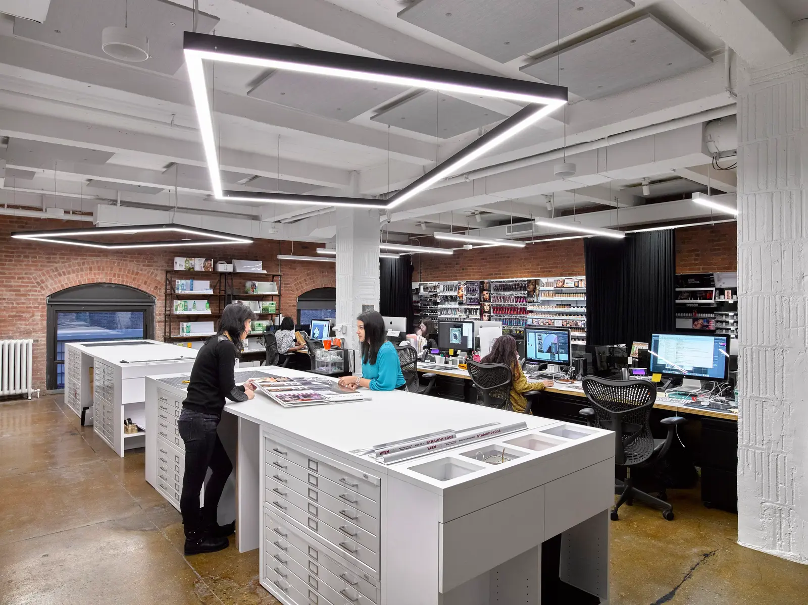 An open office design studio with exposed brick walls, geometric linear ceiling lights, white worktables with flat file drawers, and employees working at computer stations lined with cosmetics product displays.