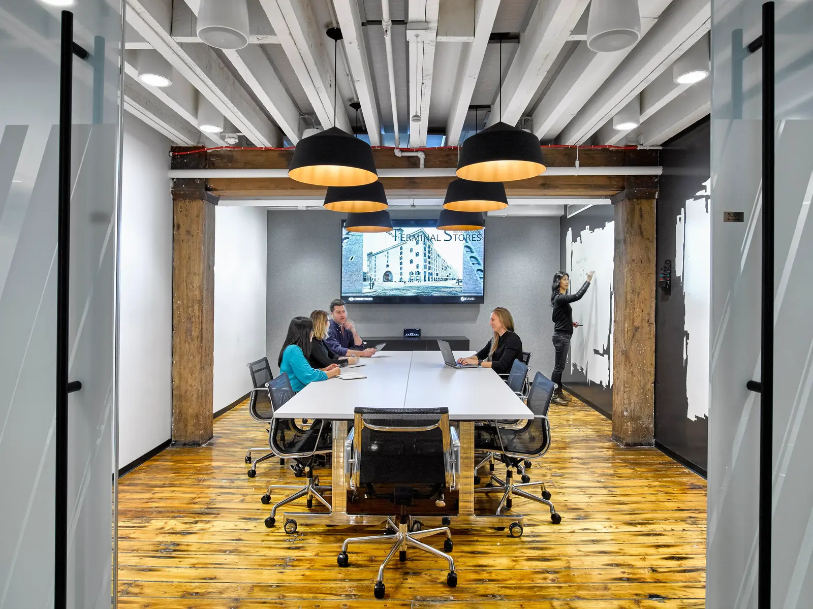 A modern conference room with exposed wood beams and black pendant lamps over a white table where colleagues meet, a wall-mounted screen displaying an image labeled 'Terminal Stores,' and a woman writing on a whiteboard in the background.