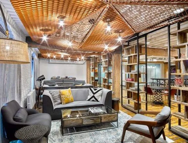 A cozy lounge area with a decorative woven wood ceiling and pendant lights, gray sofas with patterned throw pillows around a glass coffee table, and open bookshelves separating the space from a meeting area in the background.