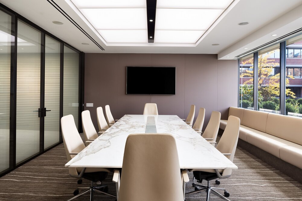Board room with conference table under bright lighting feature, next to a window.