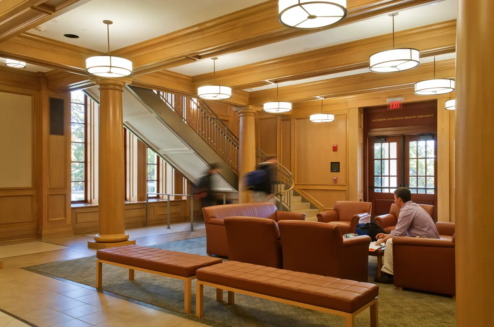 A wood-paneled lobby with tall columns and round drum light fixtures, leather armchairs and benches arranged on a patterned rug, a curved staircase in the background where two students walk up with backpacks, and a man seated reading near the exit doors.