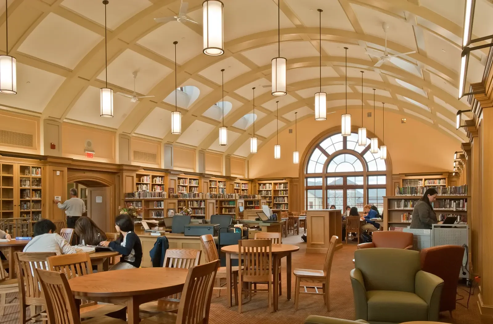Interior of a library reading room with a high vaulted, coffered ceiling and hanging cylindrical light fixtures, wooden bookshelves lining the walls, and students seated at wooden tables studying and using laptops, with a large arched window at one end.