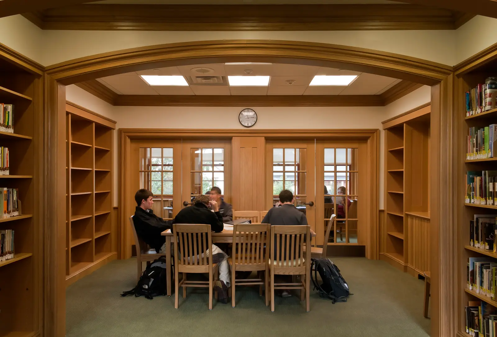 Students seated at a wooden table studying in a library alcove framed by an arched wooden doorway, with bookshelves lining both sides and glass-paned doors and a wall clock visible in the background.