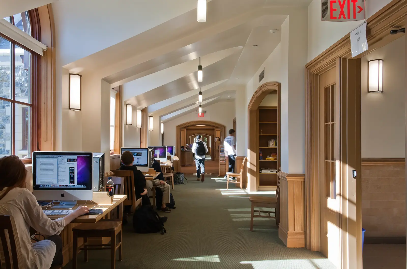 Sunlit hallway in a campus library with a vaulted wood-beamed ceiling, students seated at desks using iMac computers along the left side, and two people walking through a series of arched doorways in the background, with an illuminated 'EXIT' sign overhead.